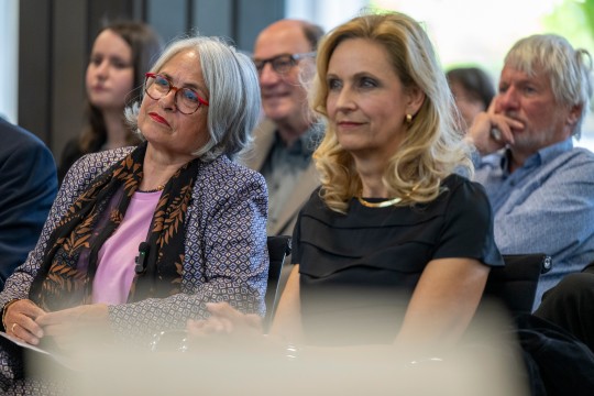 Eine Frau mit grauen schulterlangen Haaren und roter Brille sitzt neben einer blonden Frau mit schulterlangen blonden Haaren.