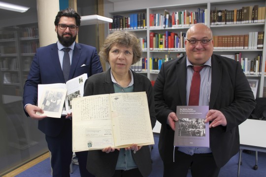 Zwei Männer und eine Frau halten in einer Bücherei offene Bücher in die Kamera.