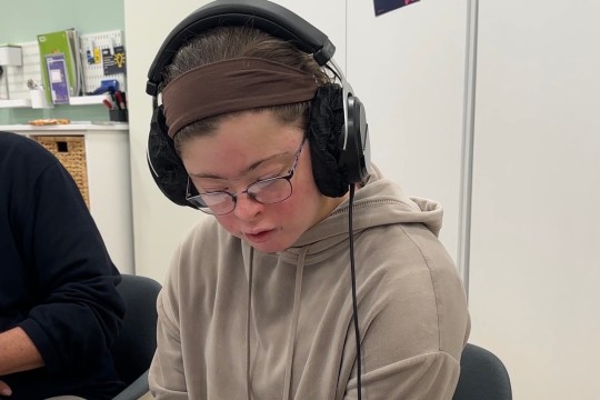 Eine Frau mit Down-Syndrom sitzt mit Kopfhörern vor einem Blatt Papier und Podcast-Aufnahmegerät