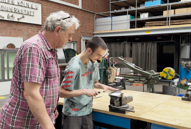Ein Mann zeigt einem Jugendlichen wie in der Metallwerstatt gefeilt wird 