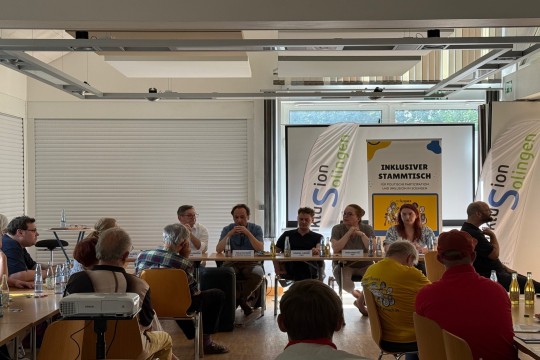 Auf dem Bild sind Menschen zu sehen. Sie sitzen vor einem Publikum.
