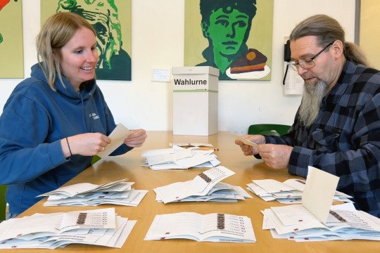 Eine Frau und ein Mann sitzen sich gegenüber an einem Tisch. Auf dem Tisch liegen viele Zettel.