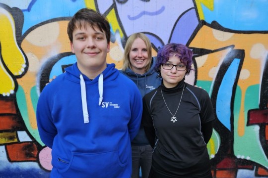 Drei Personen stehen eng zusammen und blicken lächelnd in die Kamera.